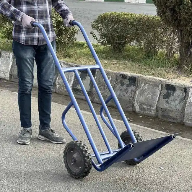 مردی با دستکش در حال حرکت دادن یک چرخ دستی حمل بار آبی رنگ در فضای باز.